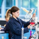 A young woman in a lab coat and safety glasses examines a volumetric flask with red liquid in a chemical technology laboratory, surrounded by colorful solutions and lab equipment