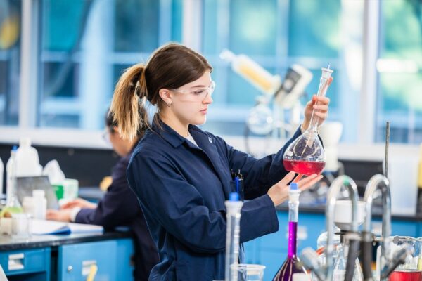 Chemical Technology: Transforming Industries and Society A young woman in a lab coat and safety glasses examines a volumetric flask with red liquid in a chemical technology laboratory, surrounded by colorful solutions and lab equipment