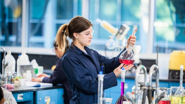 A young woman in a lab coat and safety glasses examines a volumetric flask with red liquid in a chemical technology laboratory, surrounded by colorful solutions and lab equipment