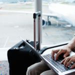 A business traveler working on a silver laptop in an airport lounge, dressed in a light blue shirt and khaki pants, with a black carry-on suitcase beside him. The background shows a blurred view of the airport tarmac with a parked airplane and ground vehicles, emphasizing a travel and productivity theme.