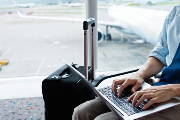 The Ultimate Guide to Lightweight Travel Laptops A business traveler working on a silver laptop in an airport lounge, dressed in a light blue shirt and khaki pants, with a black carry-on suitcase beside him. The background shows a blurred view of the airport tarmac with a parked airplane and ground vehicles, emphasizing a travel and productivity theme.