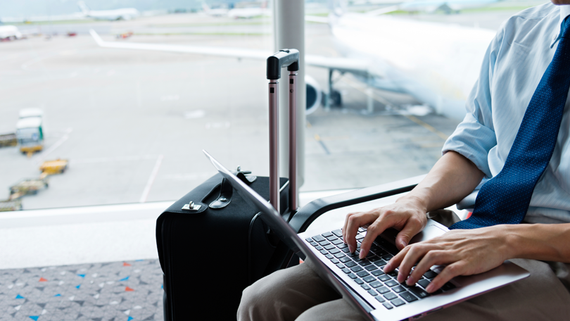 A business traveler working on a silver laptop in an airport lounge, dressed in a light blue shirt and khaki pants, with a black carry-on suitcase beside him. The background shows a blurred view of the airport tarmac with a parked airplane and ground vehicles, emphasizing a travel and productivity theme.